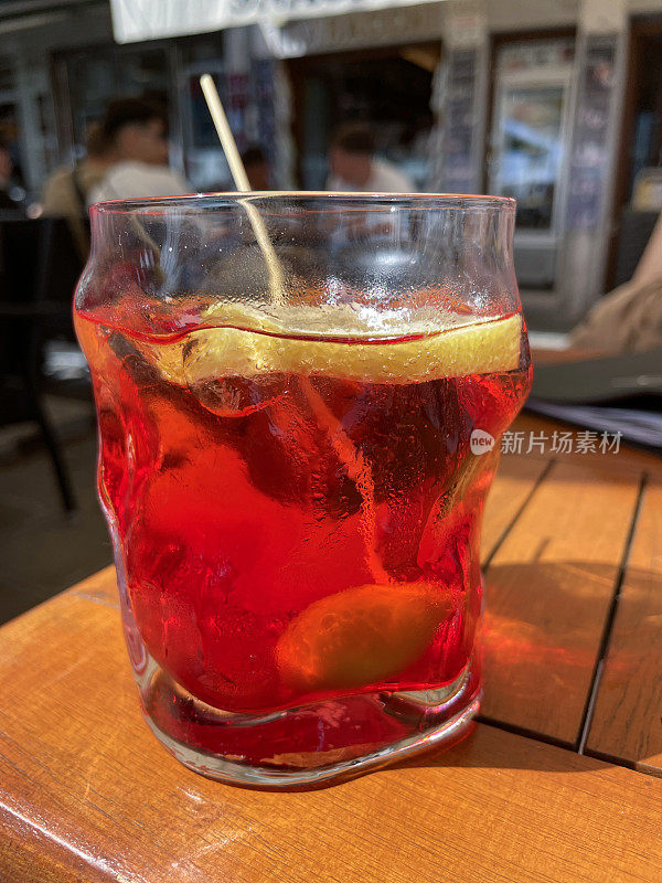 Spritz Veneziano鸡尾酒杯的特写图像，在木棒上点缀着柠檬片和橄榄，消化苦味剂，苏打水和冰块，户外用餐背景，重点放在前景